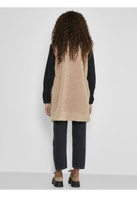 Béžová sherpa vesta s měkkou strukturou, doplněná o černé dlouhé rukávy a tmavé zkrácené kalhoty. Hnědé boty dokončují outfit.