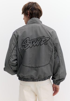 Persona che indossa una giacca a quadri bianca e nera con la scritta "Static" ricamata sulla schiena, abbinata a pantaloni avorio su uno sfondo semplice.