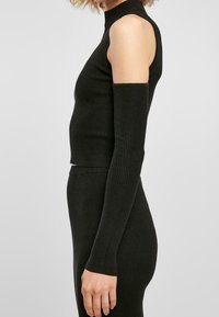 Svart ribbad topp med hög hals och öppen axeldesign, figursydda långärmar och texturerat stickat material. Tight silhuett.