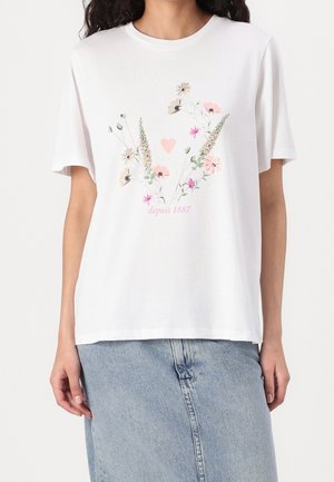 Witte T-shirt met korte mouwen met pastelbloemmotief, een klein roze hartje en tekst "depuis 1887", gedragen met een lichtblauwe spijkerrok.