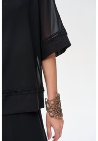 Czarny, przezroczysty top z teksturowanymi akcentami i szydełkowymi detalami na rękawach. Noszony z metalową, ażurową bransoletą na nadgarstku.