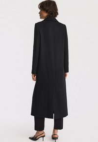 Długi czarny płaszcz wykonany z mieszanki wełny, z wysmuklonym kołnierzem, prostym krojem i rozcięciem z tyłu dla swobody ruchu. Połączony z czarnymi spodniami i obcasami.