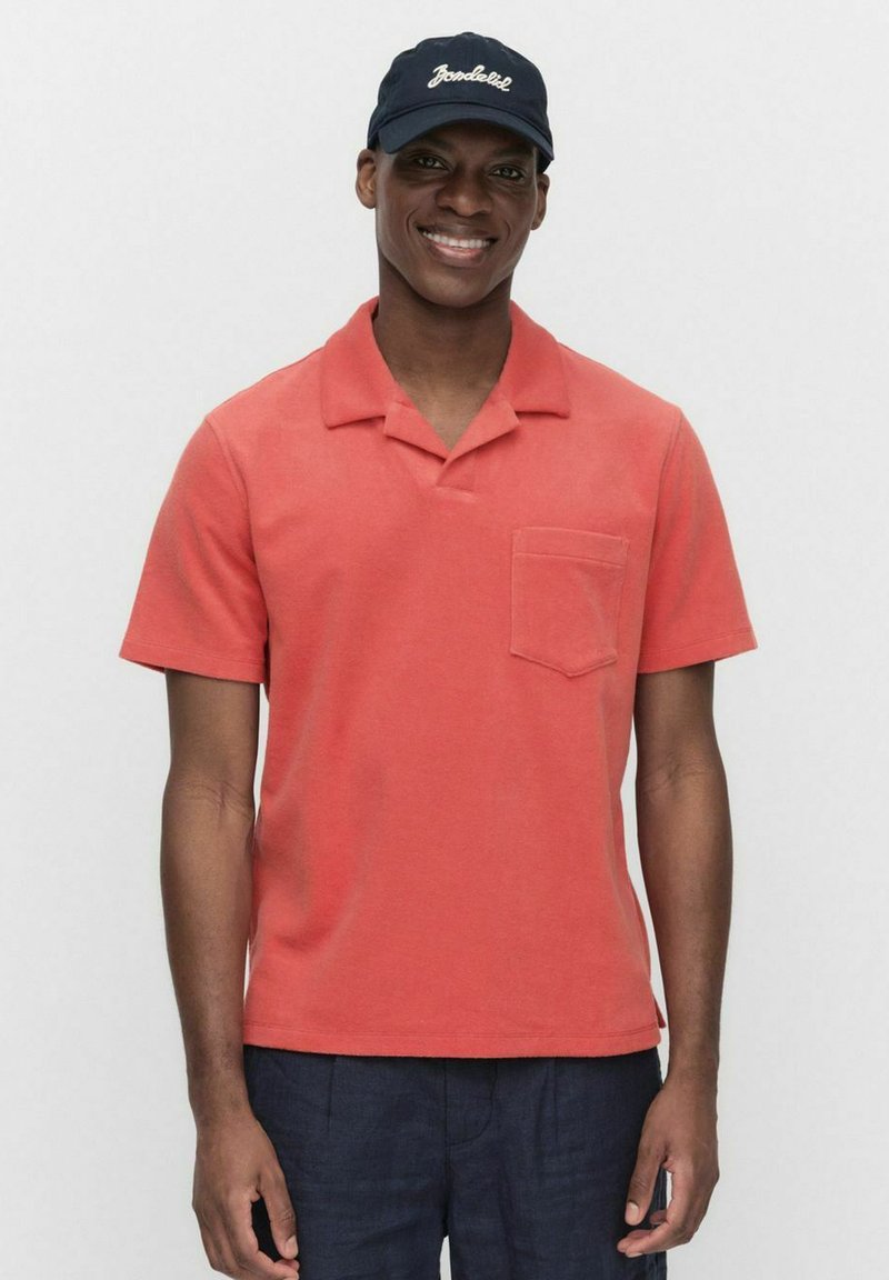 Korte mouw polo shirt in koraal katoen met een zak, platte kraag en een relaxte pasvorm. Draagt een donkere pet met witte tekst.