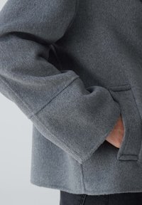Grau, strukturiere Wolljacke mit lockerem Schnitt, weiten Ärmeln und einer Seitentasche. Weiches Material mit leicht fuzzeliger Optik.