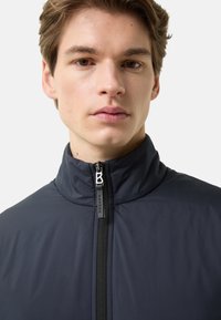 Navy blue zip-up jacket with a high collar, featuring a smooth texture, contrast zipper, and a logo tag near the zipper.