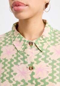 Chemise vert à motif floral rose, avec un col, des boutons et un tissu doux et texturé.
