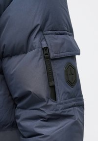 Μανίκι μπουφάν σε ναυτικό μπλε χρώμα με επένδυση, τσέπη με φερμουάρ, τσέπη με καπάκι και μαύρο εξαγωνικό έμβλημα με λογότυπο αστεριού.