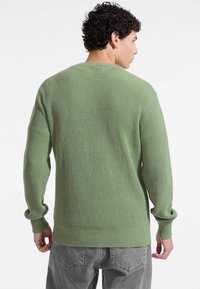 Maglione verde a costine con maniche lunghe, scollo rotondo e vestibilità rilassata. La texture è morbida, con orlo e polsini a costine.