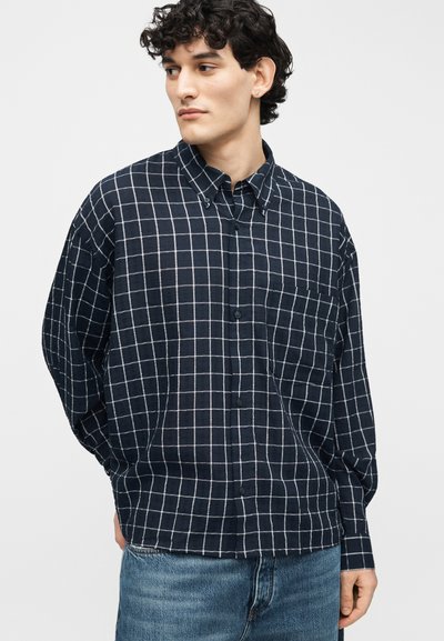 Νέος άντρας με σγουρά μαλλιά που φοράει μπλε πουκάμισο με λευκό σχέδιο πλέγματος και μπλε τζιν, κοιτάζει προς το πλάι με τα χέρια στα τσέπες.