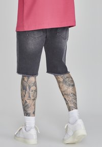 Pantalones cortos de mezclilla negra con bordes deshilachados, combinados con zapatillas blancas y calcetines blancos. Tatuajes visibles en las piernas que presentan retratos detallados y diseños.
