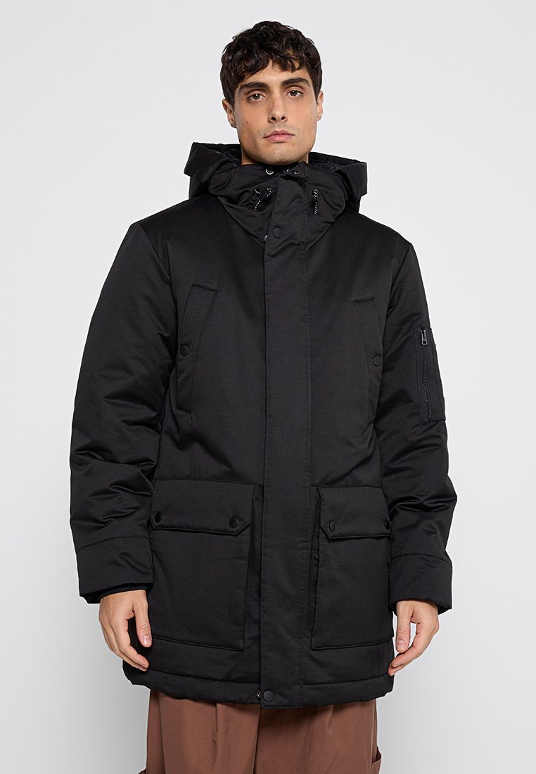 Homme portant un parka d'hiver noir à capuche avec des poches à rabat à l'avant et une poche zippée sur la manche, se tenant devant un fond uni.