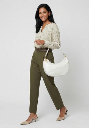 Donna con lunghi capelli scuri che indossa una camicetta beige con motivi e pantaloni verde oliva, che porta una borsa bianca con manico intrecciato, camminando e sorridendo.