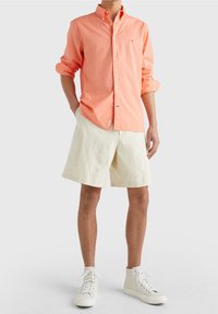 Camicia button-up di un corallo chiaro con maniche arrotolate, abbinata a pantaloni corti color crema e sneakers bianche alte. Tessuto in cotone, vestibilità casual.
