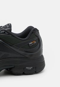 Svart atletisk sko med Cordura-tyg, texturerad ovandel, glänsande detaljer och en DMX Foam-sula för dämpning. Närbild av hälområdet.