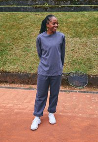 Grå langermet genser og matchende bukser laget av mykt stoff. Hvite joggesko. En tenniskølle hviler mot siden. Gress i bakgrunnen.