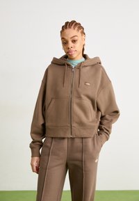 Pruun luku kaelusega lühike hoodie, mis on valmistatud pehmest kangast, sisaldab esikäe taskut ja tikitud logo. Kombineeritud sobiva pruuni dressipüksiga.