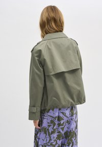 Giacca cropped verde oliva con un dettaglio arricciato sul retro, maniche lunghe con polsini abbottonati, abbinata a un indumento con stampa floreale viola sotto.