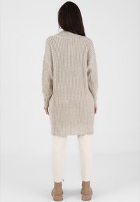 Pull oversize beige clair en tricot avec un col montant et des manches longues, motif texturé, porté avec un pantalon crème et des bottes marron.