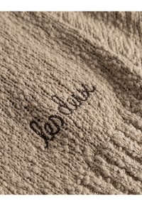 Tejido de punto beige con un patrón texturizado; presenta un guion bordado en marrón oscuro. Apariencia suave y acogedora, ideal para prendas de vestir.