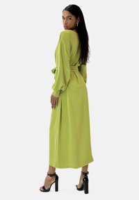 Robe à manches longues de couleur vert olive, avec une taille ceinturée, un tissu fluide et un bas légèrement effilé. Associée à des sandales à talons noires.