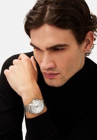 Sølv rustfrit stål ur med en hvid urskive, sorte timeindekser og et metalrem, båret på et håndled med en lys hudtone.