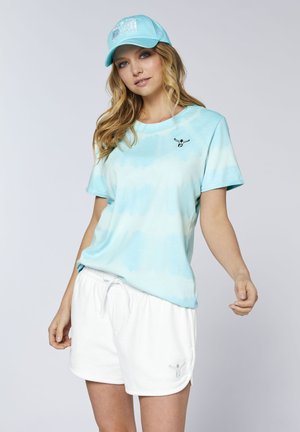 Junge Frau trägt ein hellblaues Batik-T-Shirt, weiße Shorts mit Kordelzug und eine passende blaue Baseballkappe vor einem schlichten Hintergrund.