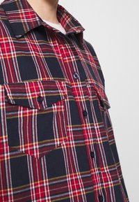 Gros plan sur une personne portant une chemise à carreaux rouge, bleu marine et blanc avec des poches poitrine à rabat, sur fond gris clair.