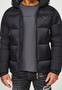 Mand iført en sort puffjakke over en grå Alessandro Zavetti Milano t-shirt og slidte sorte jeans.