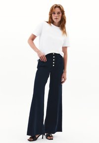 Szerokie granatowe dżinsy z pięcioma guzikami z przodu, wysoki stan i postrzępione wykończenie. W połączeniu z gładką białą koszulką z krótkim rękawem.