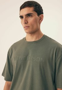 Oliwkowy t-shirt z krótkim rękawem wykonany z bawełny o teksturowanym wykończeniu. Z przodu znajduje się dyskretny, wytłoczony logo "HENDERSON."