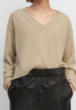 Pull en maille beige à col en V associé à un pantalon en cuir noir taille haute, mains dans les poches, présenté du cou aux hanches.