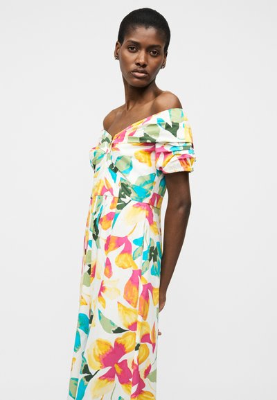 Sieviete īsiem matiem baltā kleitā ar noslīdošām pleciem un krāsainu lielu ziedu rakstu, stāv pret vienkāršu gaišu fonu.