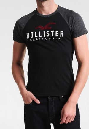 Mężczyzna ubrany w czarno-szary T-shirt Hollister California z krótkim rękawem i ciemne dżinsy, z ręką w kieszeni, na jednolitym tle.