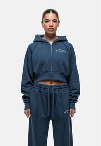 Afgekorte marineblauwe hoodie met rits en capuchon, heeft een zachte textuur en geborduurd logo. Gepaard met bijpassende joggingbroek met aantrekkoord.