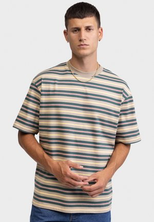 Νέος άνδρας που φοράει κοντομάνικο πουκάμισο με ρίγες σε μπεζ, πράσινο, κίτρινο και κόκκινο, χρυσή αλυσίδα και μπλε τζιν, στέκεται με τα χέρια δεμένα.