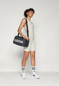 Must küllane Adidas duffel kott valge logo, ristkülikukujuline, kangasmaterjal, mida kannab kerge halli sportliku moe ja valgete kingadega inimene.