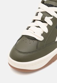 Sneaker verde oliva con tomaia in pelle liscia, lacci bianchi morbidi e suola in gomma. Presenta perforazioni sulla punta e dettagli cuciti.