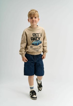 Beige kapucnis pulóver grafikus nyomattal, amelyen egy kék autó látható, kék farmer rövidnadrággal és fekete sportcipővel, fehér és kék csíkos zoknival párosítva.