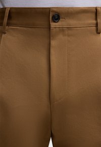 Primo piano di pantaloni marroni con un bottone nero e passanti per cintura visibili sulla vita.