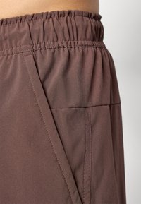 Calções castanhos feitos de um tecido leve e suave. Apresentam um cós elástico e um bolso lateral, com costuras limpas visíveis ao longo das costuras.