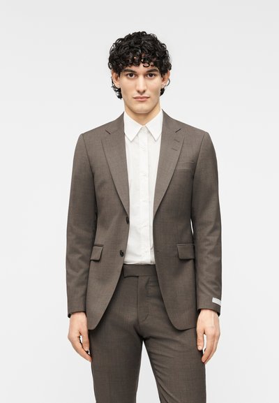 Καφέ κοστούμι ραμμένο στα μέτρα με στενή γραμμή, διαθέτει γιακά με εγκοπή, δύο μπροστινές τσέπες και υφή υφάσματος. Συμπληρώνεται με λευκό πουκάμισο από κάτω.