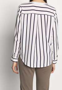 Chemise blanche à manches longues avec des rayures verticales bleu marine, ourlet arrondi, pli d'aisance au dos et poignets à boutons. Associée à un pantalon beige.