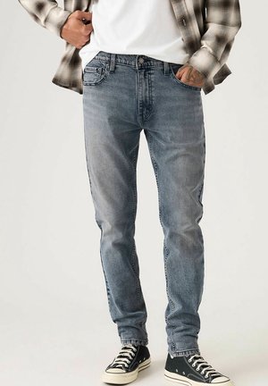 Homme portant un jean bleu délavé, des baskets noires et blanches, un t-shirt blanc et une chemise à carreaux beige les mains dans les poches.
