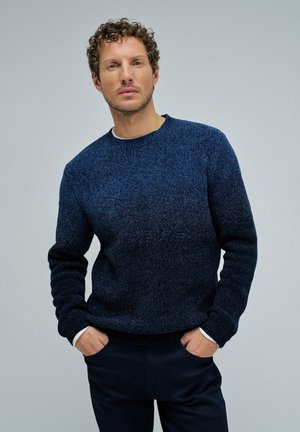 Hombre con cabello rizado que lleva un suéter azul texturizado y pantalones oscuros, de pie con las manos en los bolsillos frente a un fondo gris liso.