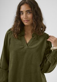 Mlada žena z valovitimi rjavimi lasmi, ki nosi olivno zeleno žametno majico, z roko prilagaja Rokav, nevtralna ozadje.