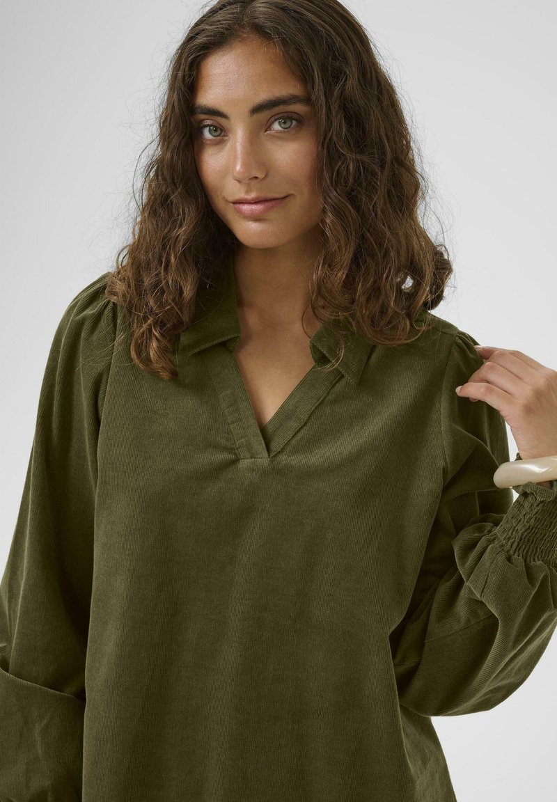 Mlada žena z valovitimi rjavimi lasmi, ki nosi olivno zeleno žametno majico, z roko prilagaja Rokav, nevtralna ozadje.