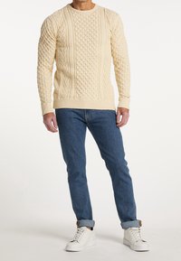 Cremfarbener Strickpullover mit strukturiertem Zopfmuster, Rundhalsausschnitt und gerippten Bündchen, kombiniert mit blauen Jeans und weißen Sneakers.
