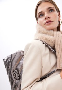 Mladá žena v krémovém kabátu a béžovém šátku se postaví s kovovým batohem před bílým pozadím a dívá se stranou.