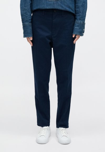 Jūras zils kokvilnas bikses ar brīvu piegriezumu, kurām ir pogu jostasvieta un kabatas, valkātas kopā ar baltām sporta kurpēm uz vienkārša fona.