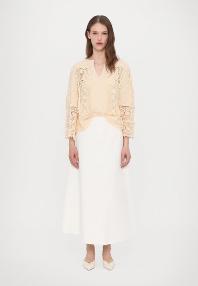 Kardet, blonde bluse i lyse beige med V-hals og lange ærmer, parret med en fodlang hvid nederdel og spidse beige sko.